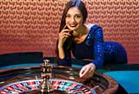 Crupier real en un casino online en vivo posando al lado de una ruleta.