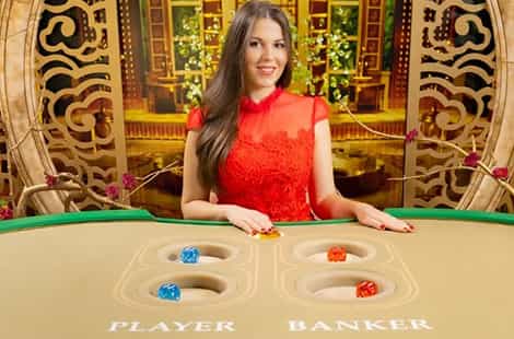 Croupier pelirroja de pelo largo en un salón de juego, sentada de lado junto a una ruleta, de lado, con un jarrón de pie con flores y otra mesa de juego al fondo.