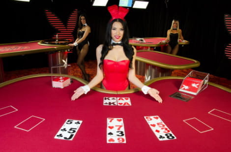 Mesas de blackjack Playboy con cuatro mesas. En primer plano, aparece una mano de blackjack con una crupier morena presentando la mesa con las palmas de las manos hacia arriba y ataviada con un body rojo, pajarita y orejas de conejita. En el fondo aparecen otras dos conejitas de Playboy apoyadas en otras dos mesas de casino.