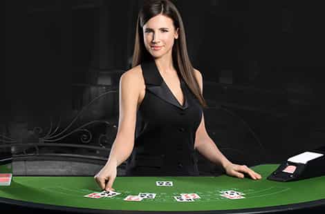 Crupier sonriente ataviada con un traje de chaqueta y una camisa blanca con los brazos apoyados sobre una ruleta, en una sala con un fondo verde.