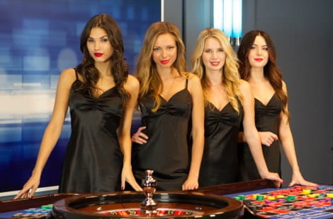 Cuatro mujeres crupieres ataviadas con vestidos negros similares, de pie ante una mesa con una ruleta y un tapete, en un estudio con un fondo de color azul y blanco.