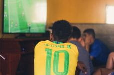 Un joven ve un partido de fútbol por televisión con una camiseta de la selección brasileña de fútbol con el nombre de Neymar.