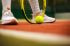 Pies de un tenista junto a una raqueta apoyada en la pista y una pelota.