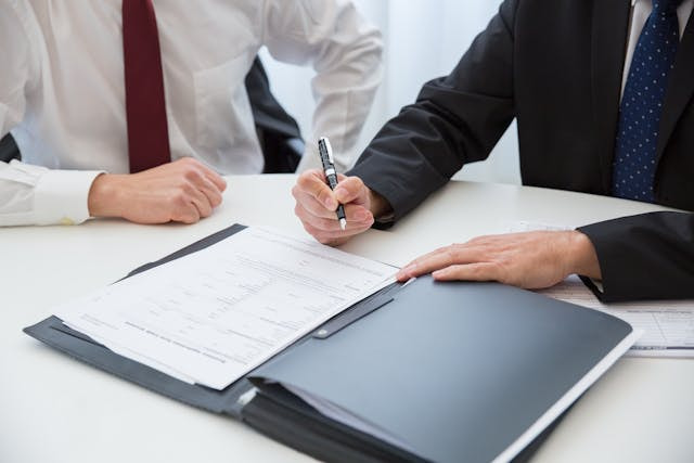 Dos empresarios en una mesa mientras uno de ellos firma un documento en un dosier.