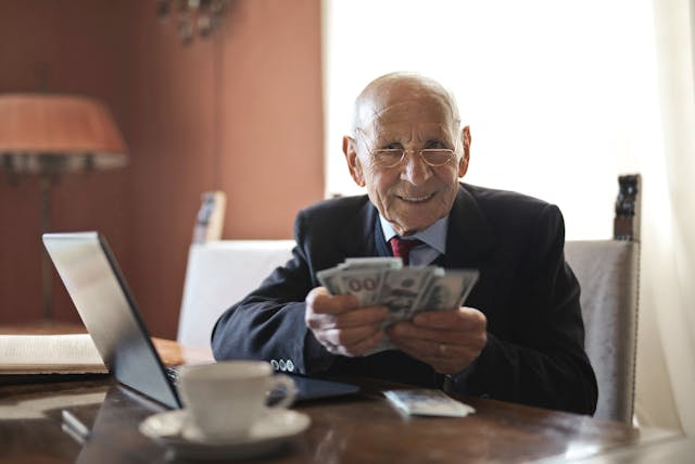 Adulto mayor sentado en su escritorio frente a su portátil con varios billetes en sus manos.