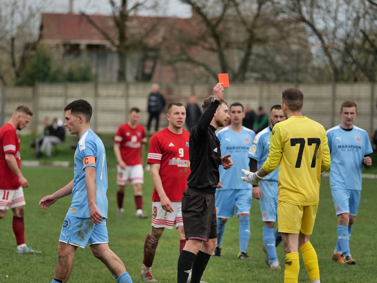 Un árbitro sacando una tarjeta rosa en mitad de un partido de fútbol rodeado por jugadores de ambos equipos.