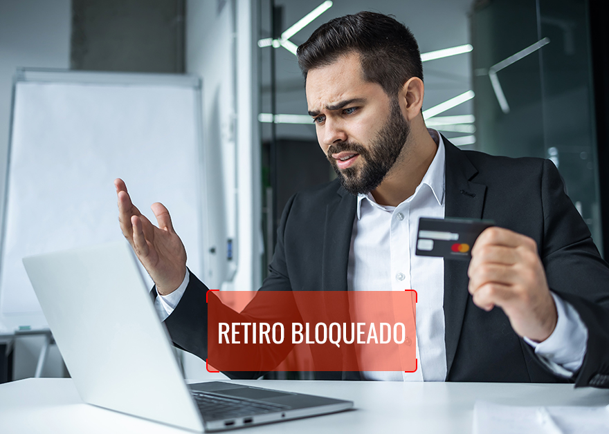Hombre frustrado con tarjeta de crédito frente a un portátil con el mensaje “Retiro bloqueado”.