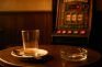 Mesa de bar con un vaso vacío, un plato con una cucharilla y un cenicero con una colilla, con una máquina tragaperras desenfocada al fondo.