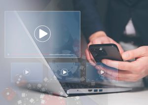 Persona usando un portátil y un móvil con iconos de reproducción superpuestos, representando contenido en vídeo.