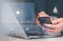 Persona usando un portátil y un móvil con iconos de reproducción superpuestos, representando contenido en vídeo.