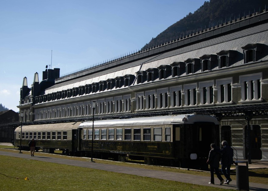 Vista exterior de la estación de Canfranc con vagones históricos frente al edificio y montañas al fondo.