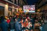 Multitud de aficionados viendo un partido en pantalla gigante en una plaza con bar.