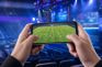 Manos sujetando un móvil con un partido de fútbol virtual en un pabellón de esports iluminado