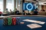 Fichas de póker apiladas y cartas sobre una mesa de torneo con el reloj de niveles al fondo