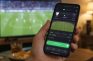 Mano con un móvil que muestra una apuesta en directo frente a un partido en la televisión