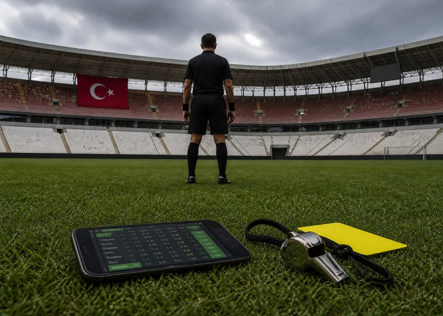 Árbitro en un estadio turco vacío, con un móvil de apuestas y un silbato en el césped